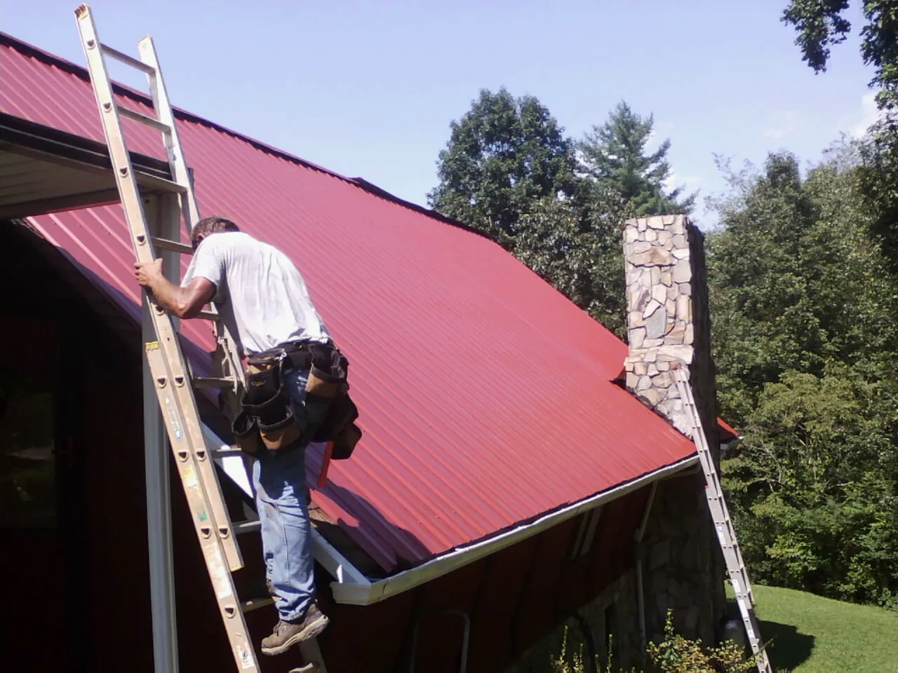Roofer on a ladder beside a red metal roof and stone chimney — Soffit & Fascia Repair by Roofing Brothers in Sikeston